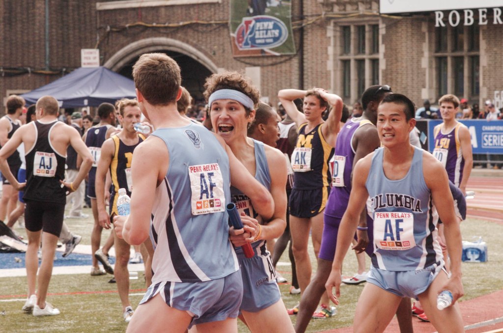 A runner in a headband and Columbia track and field singlet and shorts celebrates with two other columbia runners